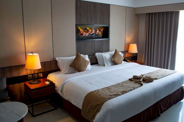 A cozy hotel room features a king-sized bed, two bedside tables with lamps, a wall-mounted fireplace, and a window with curtains.