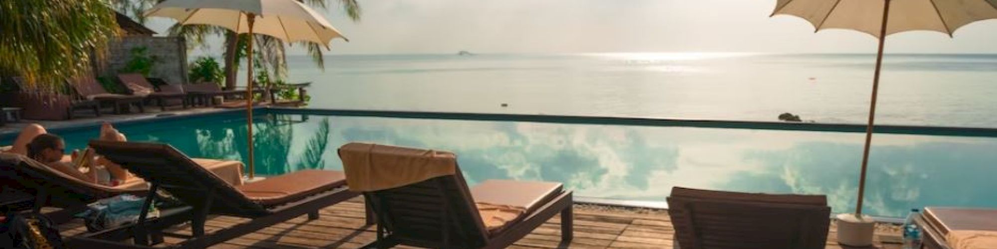 The image shows a serene poolside with lounge chairs, umbrellas, and an infinity pool overlooking a calm sea, under a partly cloudy sky.