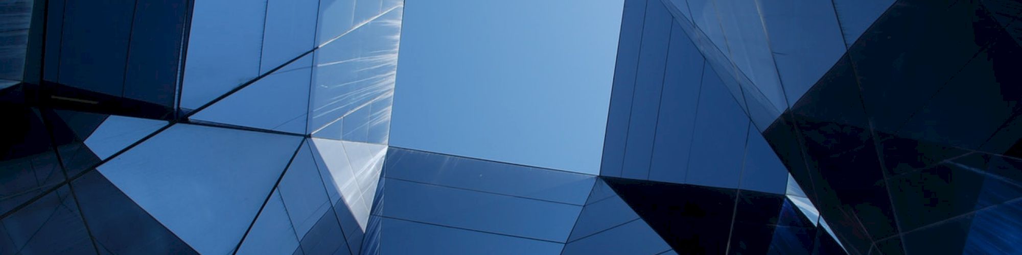 The image shows an upward view of a modern building with mirrored glass panels reflecting the sky, creating a symmetrical geometric pattern.
