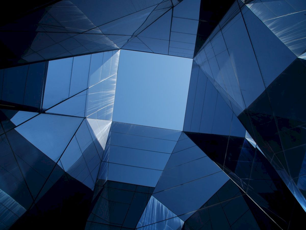 The image shows a geometric, modern architectural structure with glass panels reflecting the sky, creating a symmetrical, abstract pattern.