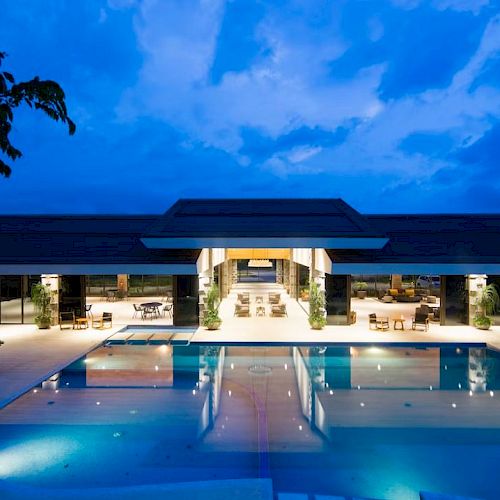 An illuminated poolside view of a modern building with large open spaces and seating areas, beautifully lit against a twilight sky ending the sentence.