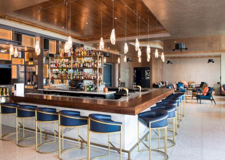 The image shows an upscale bar with a wooden counter, blue bar stools, hanging lights, and a well-stocked shelf of bottles. There's also seating in the background.