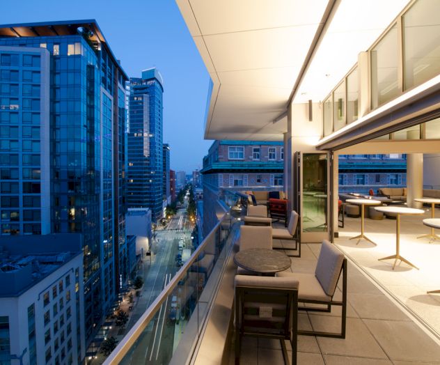 A modern urban balcony with outdoor seating overlooks a city street lined with high-rise buildings, captured during twilight.