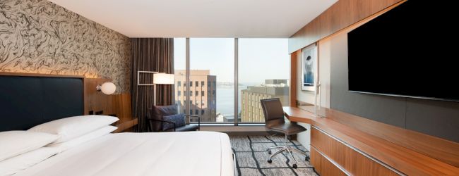 A modern hotel room featuring a king-size bed, large TV, desk, and chair with a view of city buildings and harbor through the window.