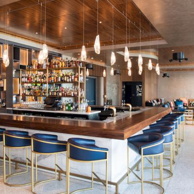 This image shows a modern bar with a wooden counter, blue bar stools, a well-stocked liquor shelf, and pendant lights, with seating areas in the background.
