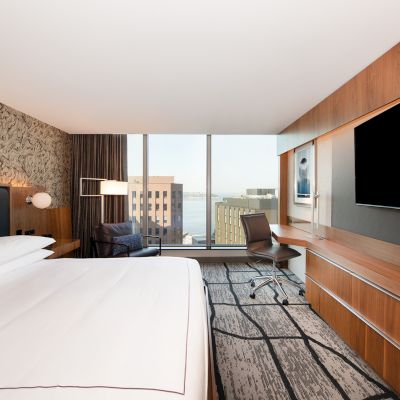 A modern hotel room with a large bed, desk, chair, and flat-screen TV. The window offers a city view with buildings and a glimpse of the water outside.