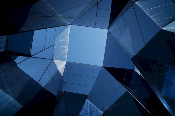 The image shows a geometric architectural structure with reflective glass panels, creating a view looking up towards the sky.
