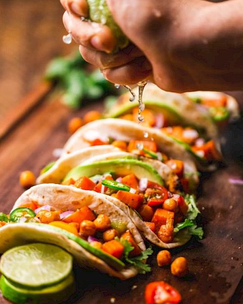 A hand is squeezing lime juice onto a row of tacos filled with chickpeas, avocado slices, cilantro, and diced vegetables arranged on a wooden board.