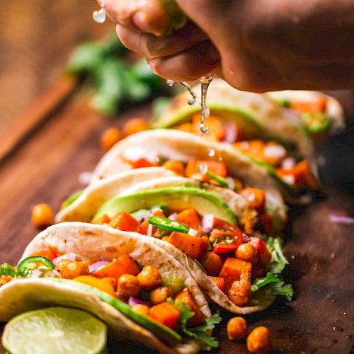 A hand is squeezing lime juice onto a row of tacos filled with chickpeas, avocado slices, cilantro, and diced vegetables arranged on a wooden board.