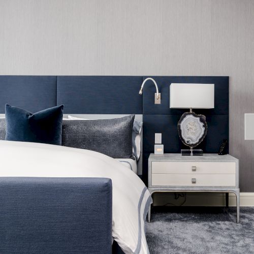 A modern bedroom with a blue and white color scheme features a bed, blue headboard, pillows, nightstand, lamp, decorative item, and wall-mounted reading light.