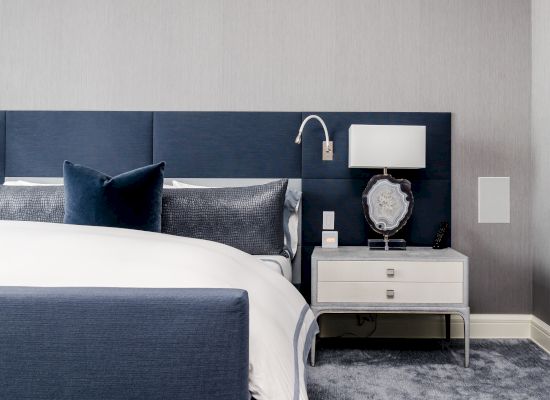 A modern bedroom with a blue and white color scheme features a bed, blue headboard, pillows, nightstand, lamp, decorative item, and wall-mounted reading light.