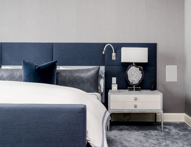 A modern bedroom with a blue and white color scheme features a bed, blue headboard, pillows, nightstand, lamp, decorative item, and wall-mounted reading light.