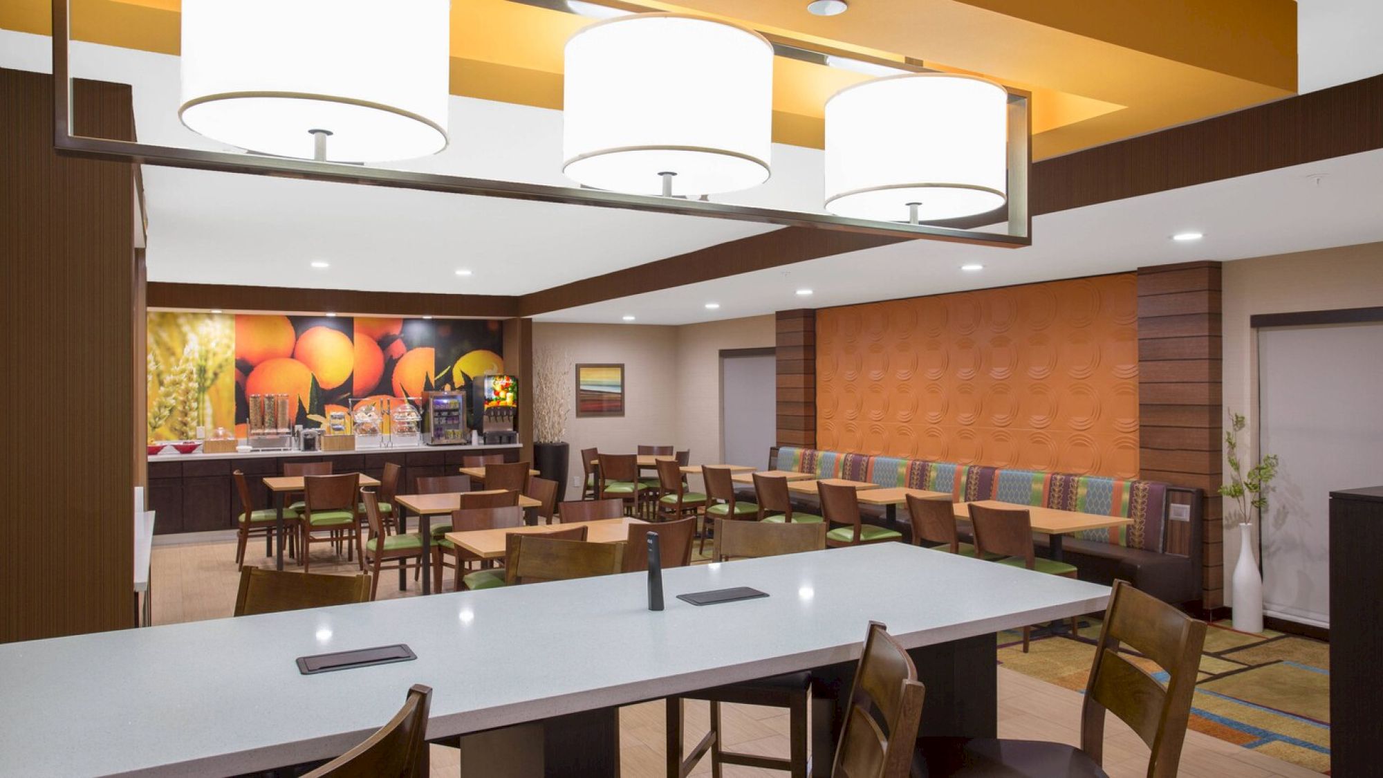 A spacious dining area features modern lighting, a variety of seating options, and a food service counter with a colorful mural in the background.