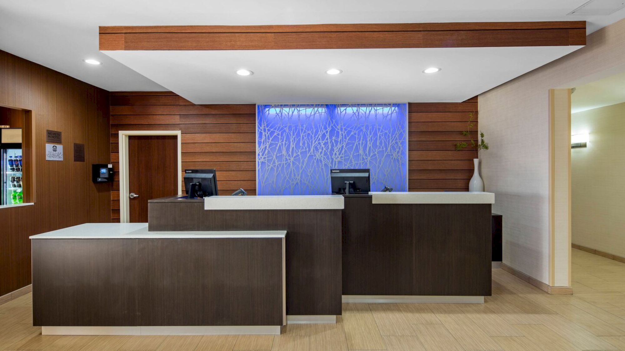This image shows a modern hotel reception area with two desks, computers, a vase, and backlit blue artwork on the wall behind.