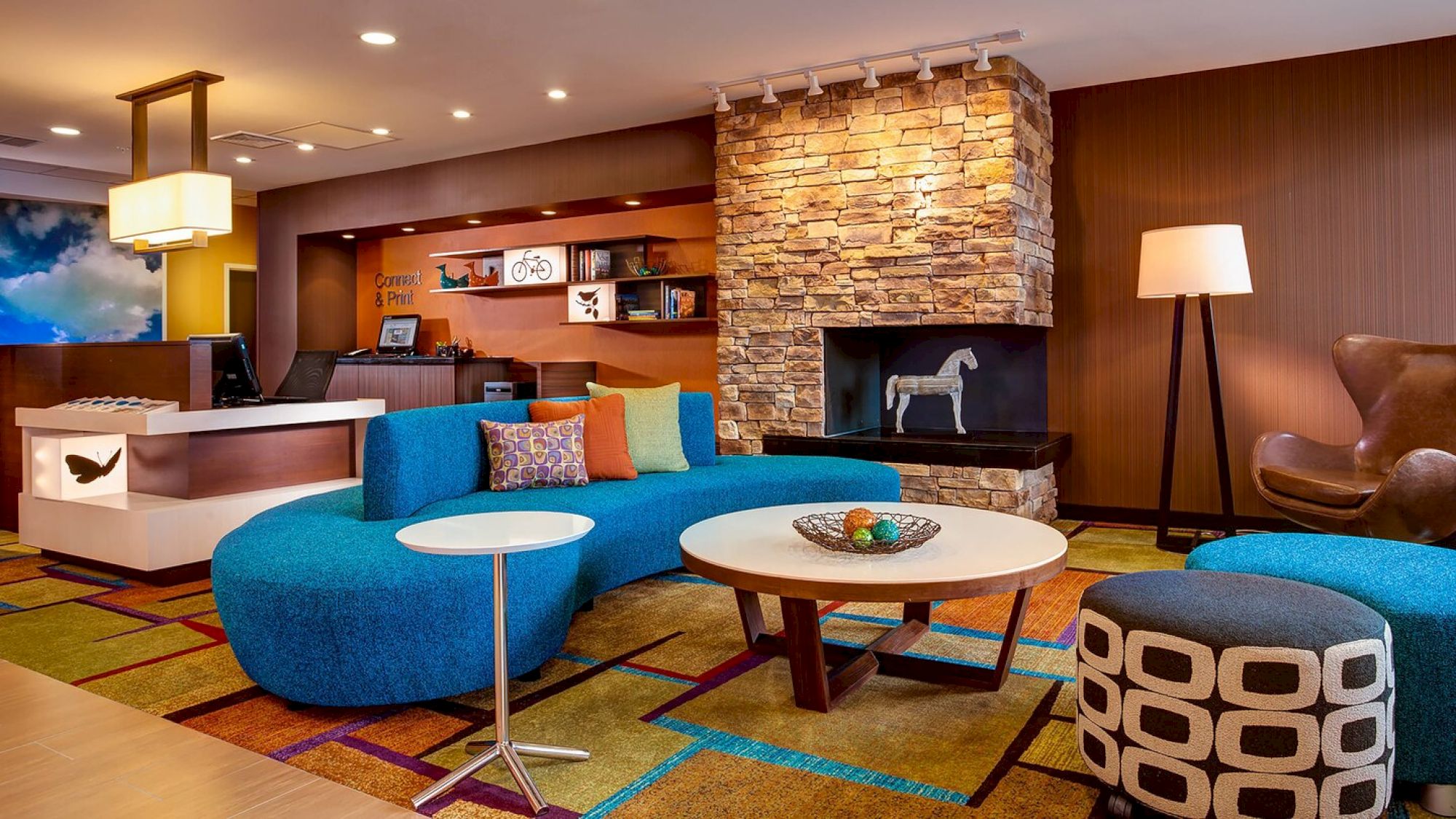 Modern hotel lobby with a blue curved sofa, colorful cushions, a stone fireplace, abstract décor, and contemporary furniture and lighting fixtures.