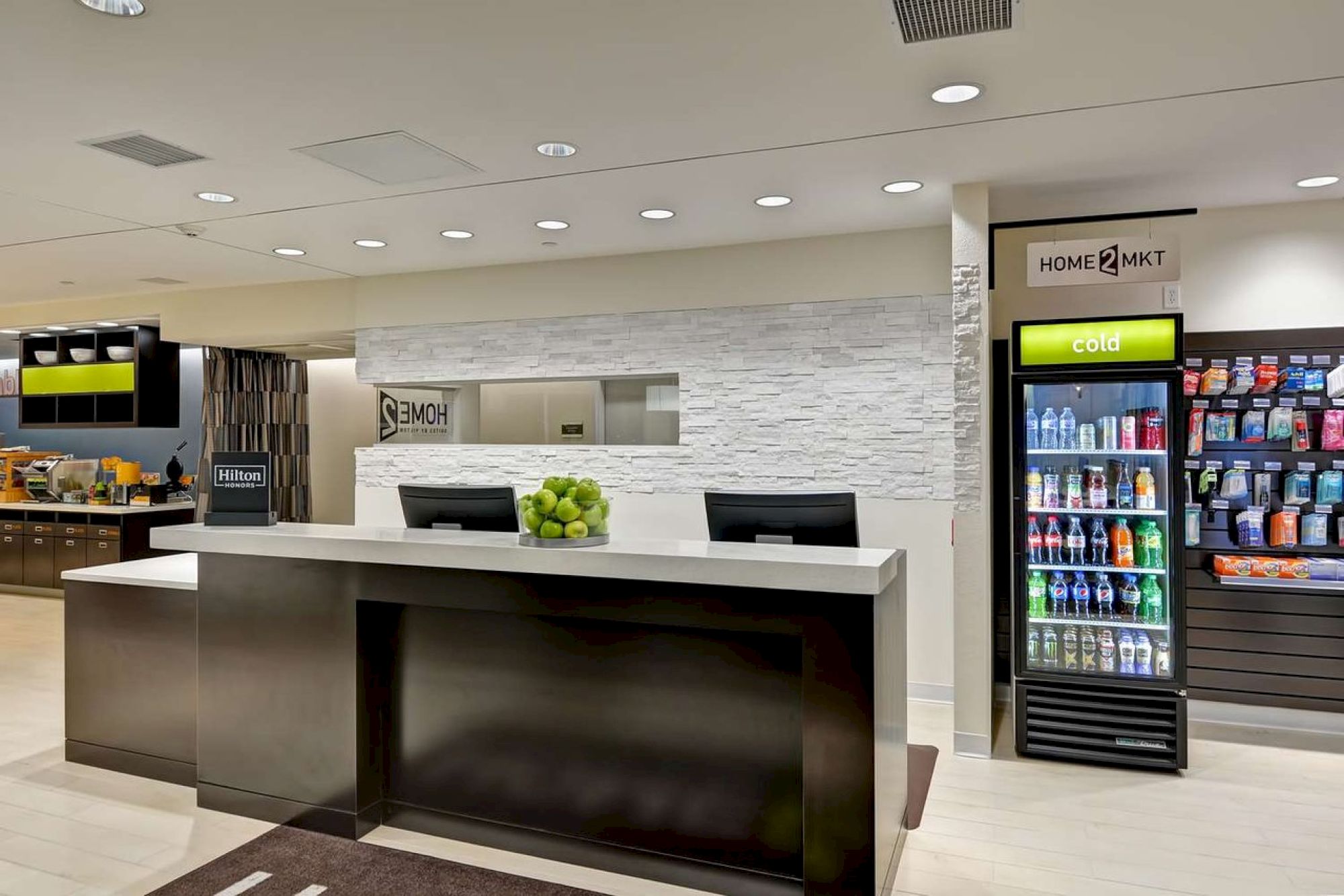 This image shows a modern hotel reception with two black counters, a bowl of green apples, a drink/snack vending machine, and a sign that reads 