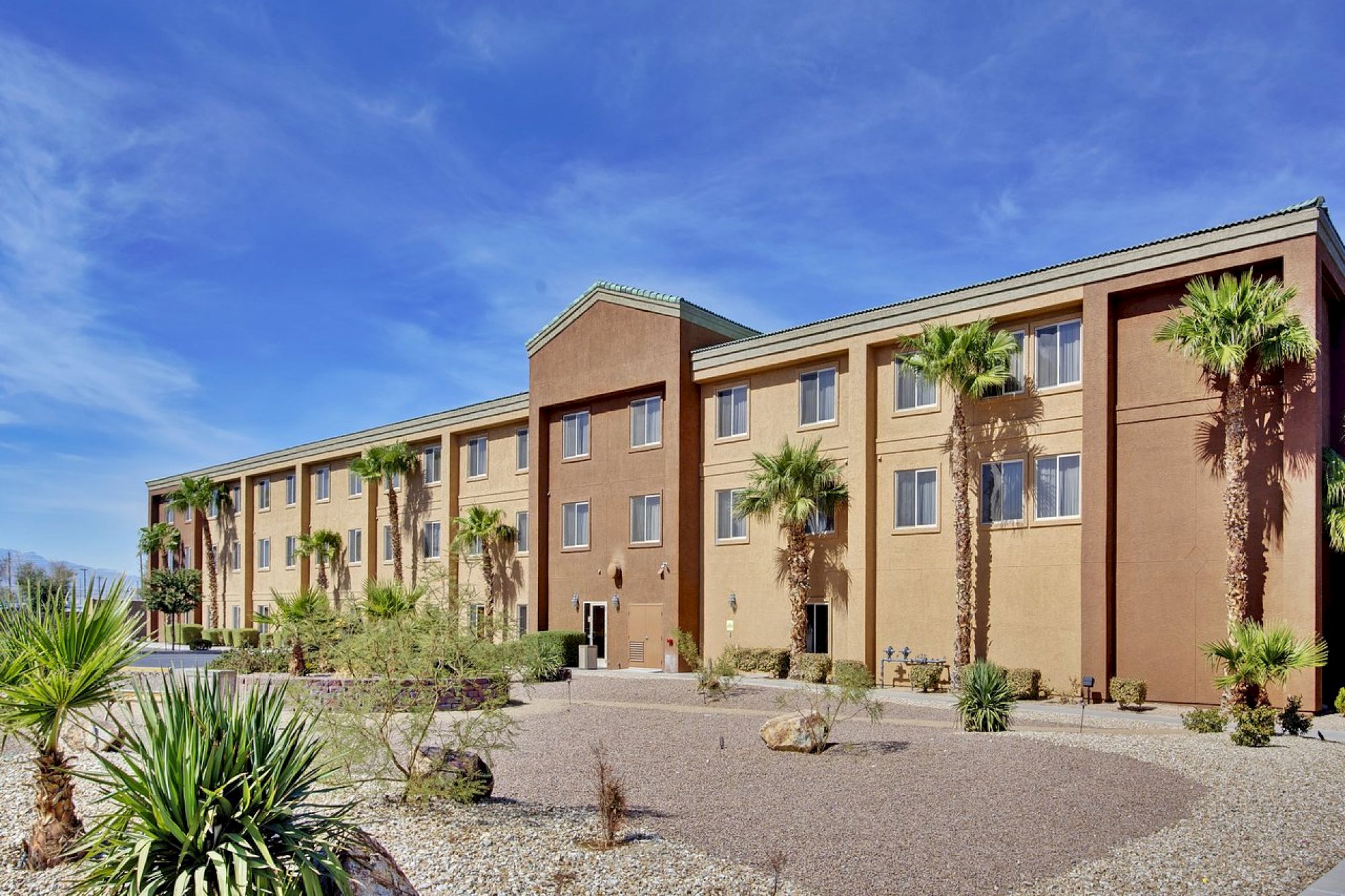 A three-story beige building with palm trees and desert landscaping in front, under a clear blue sky.