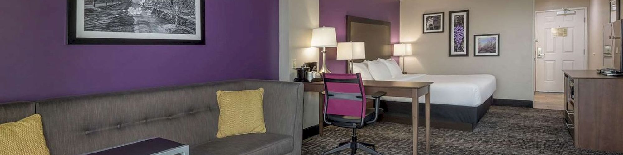 A modern hotel room features a seating area, a desk, a bed, and framed artworks on purple walls, providing a cozy and inviting ambiance.