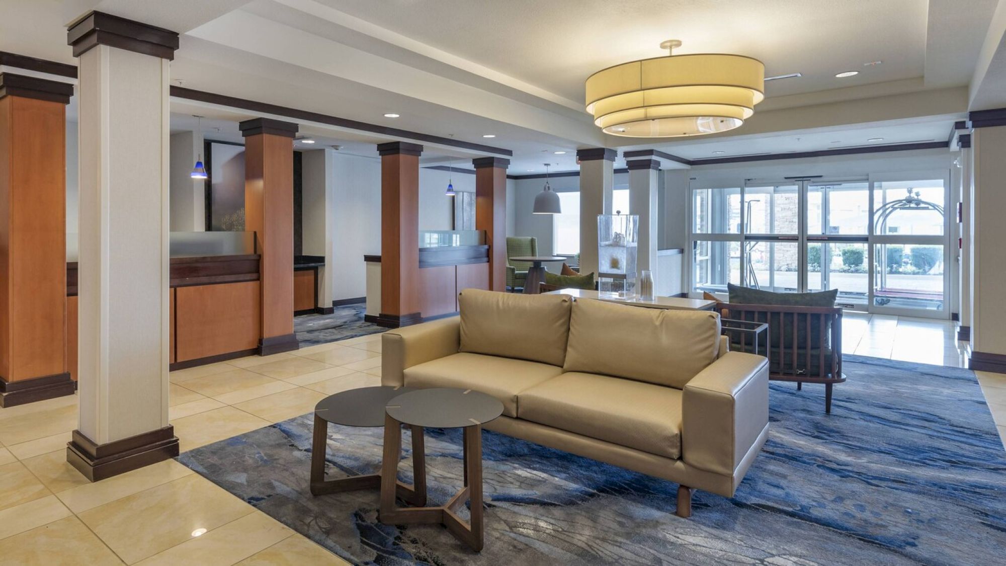 A modern hotel lobby with a beige sofa, small tables, reception area, glass entrance doors, and contemporary decor.