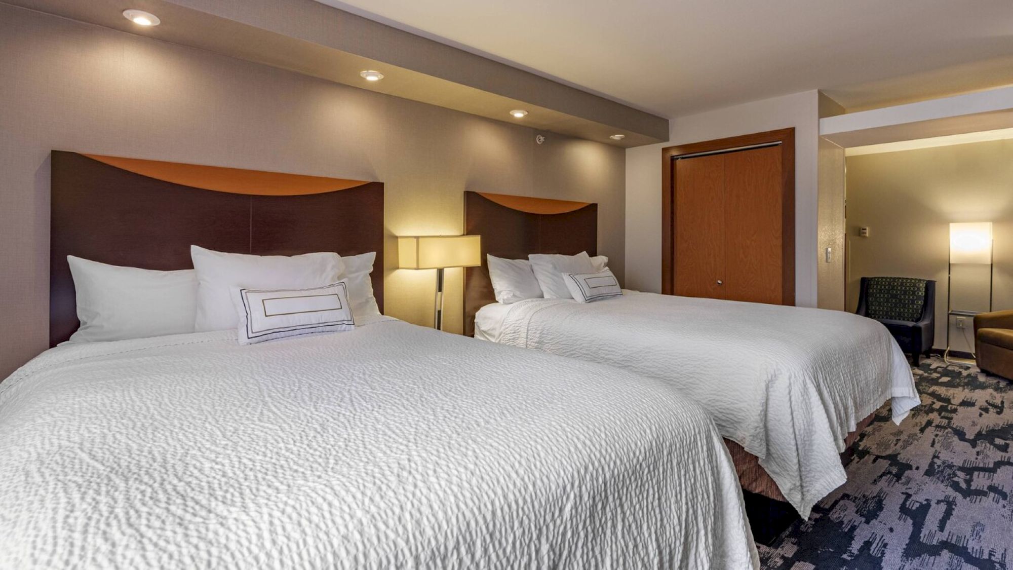 The image shows a hotel room with two large beds, wall-mounted headboards, white bedding, a lamp between the beds, and modern decor.