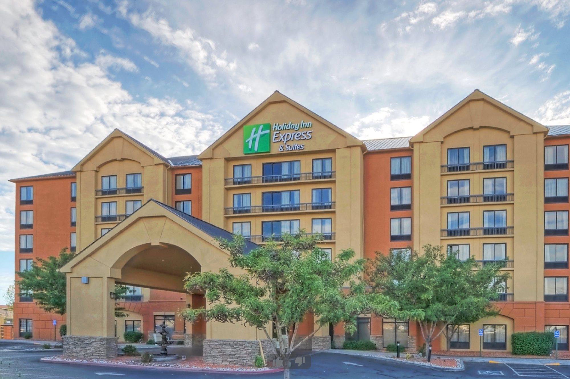 The image shows the exterior of a Holiday Inn Express & Suites hotel with a well-maintained entrance, trees, and a partly cloudy sky in the background.