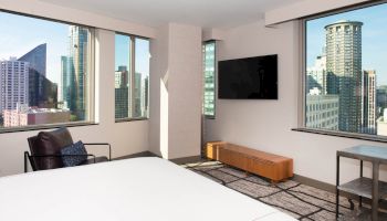 A modern hotel room with city views, a mounted TV, chair, and minimal décor, showcasing large windows with an urban skyscraper backdrop.