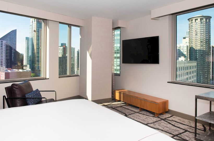 A modern hotel room with city views, a mounted TV, chair, and minimal décor, showcasing large windows with an urban skyscraper backdrop.