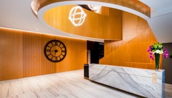 The image shows a modern lobby with a curved wooden staircase, a marble reception desk, a wall clock, and decorative lights and flowers.