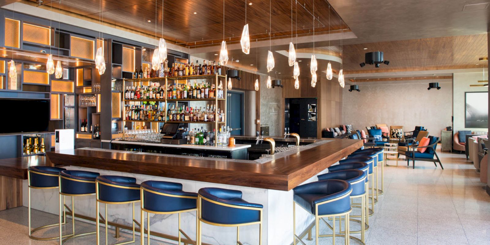 A stylish modern bar with a stocked liquor shelf, blue cushioned bar stools, overhead pendant lights, and a cozy seating area in the background.