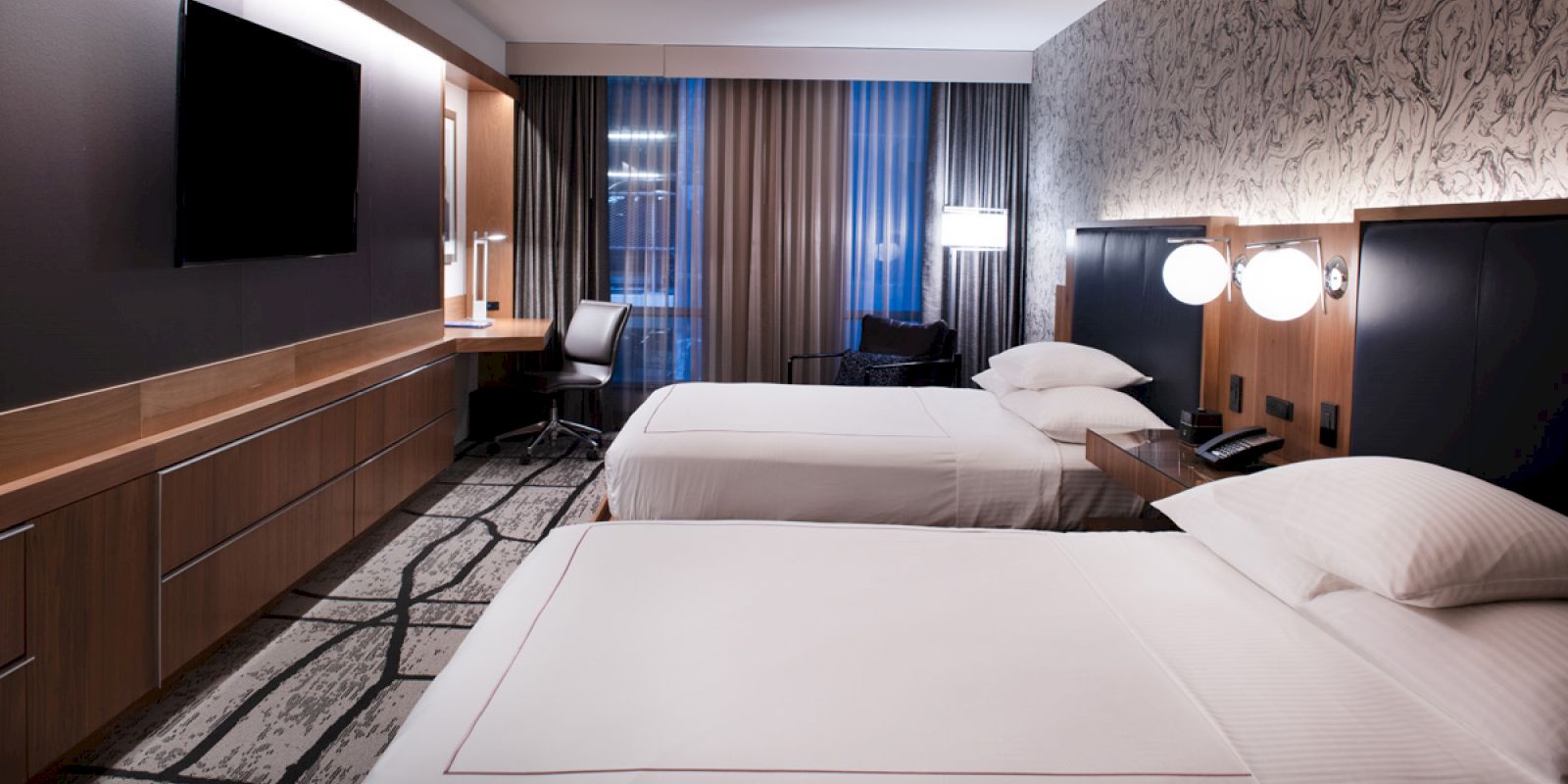 This image shows a modern hotel room with two beds, a wall-mounted TV, a desk with a chair, and ambient lighting.