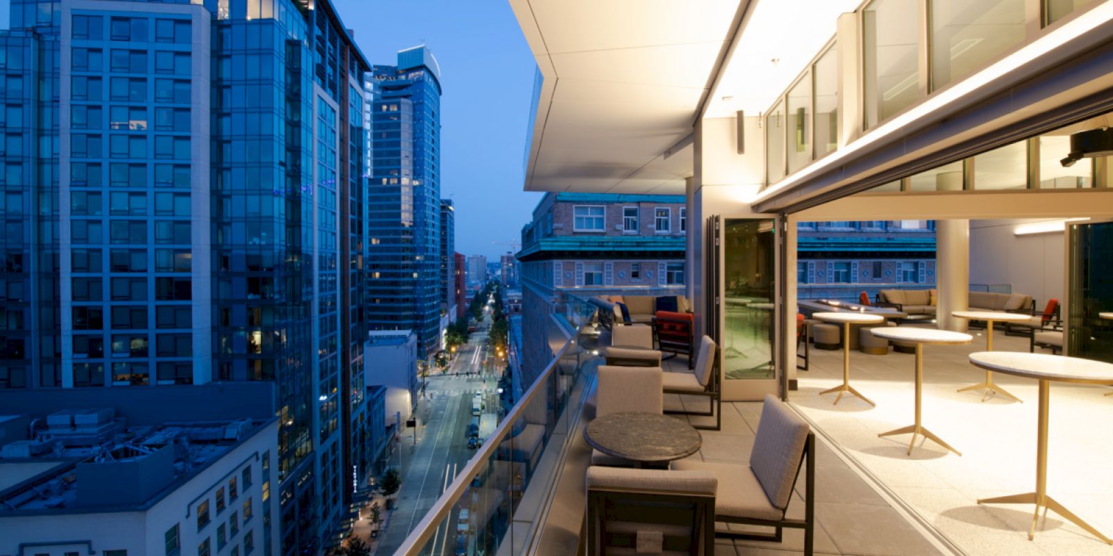 The image shows a modern cityscape at dusk, with a balcony featuring seating and tables overlooking tall buildings and a street.