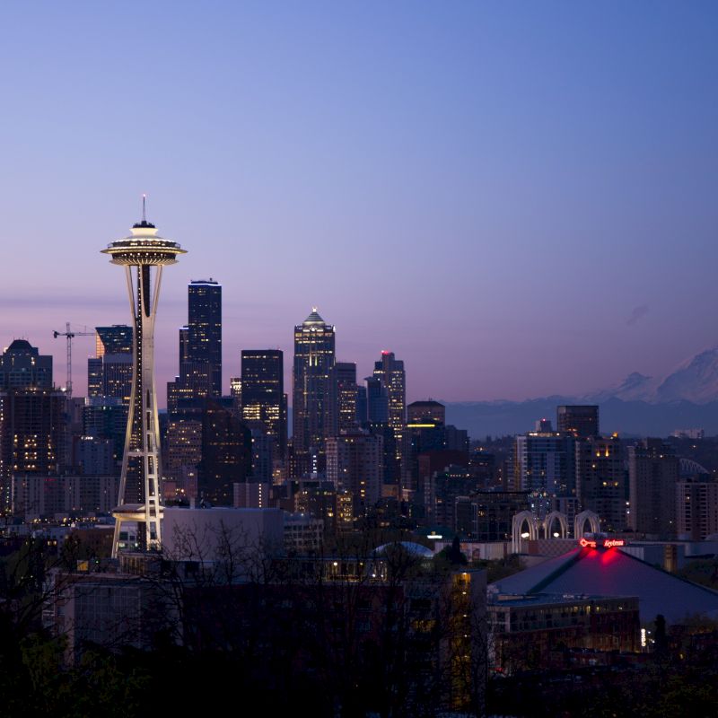 This image shows the Seattle skyline at dusk, with the Space Needle prominently visible and Mount Rainier in the background, under a clear sky.