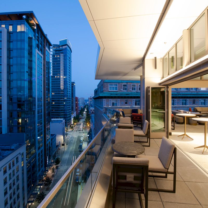 A modern balcony view of a cityscape at dusk, featuring buildings, a street below, and a well-furnished outdoor seating area.