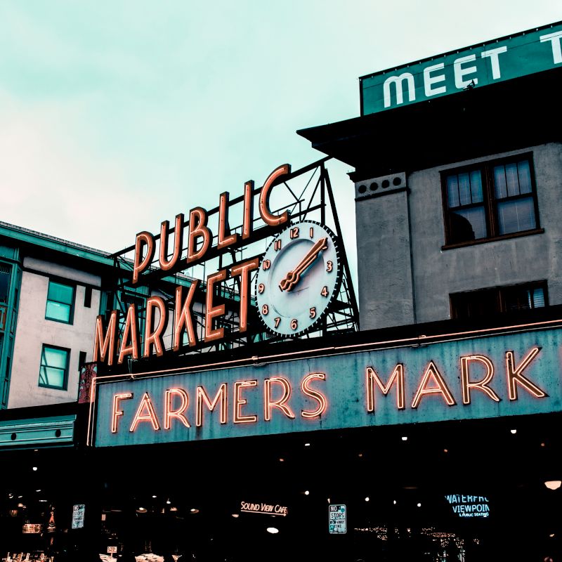 This image shows a sign for a public farmers market with a clock on a building, located in an urban area with additional signage and buildings visible.