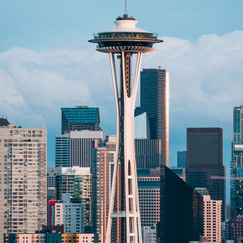 The image shows the Space Needle in Seattle, surrounded by various tall buildings and a cloudy sky in the background.