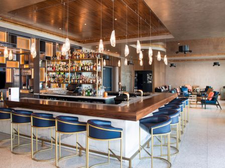 A modern bar with a wooden countertop, blue bar stools, hanging pendant lights, and a well-stocked liquor shelf in a spacious, stylish setting.