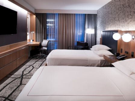 The image shows a modern hotel room with two neatly made beds, a desk, a flat-screen TV, and ambient lighting, creating a cozy atmosphere.