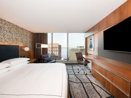 This image shows a modern hotel room with a large bed, TV, desk, chair, and windows with a cityscape view.