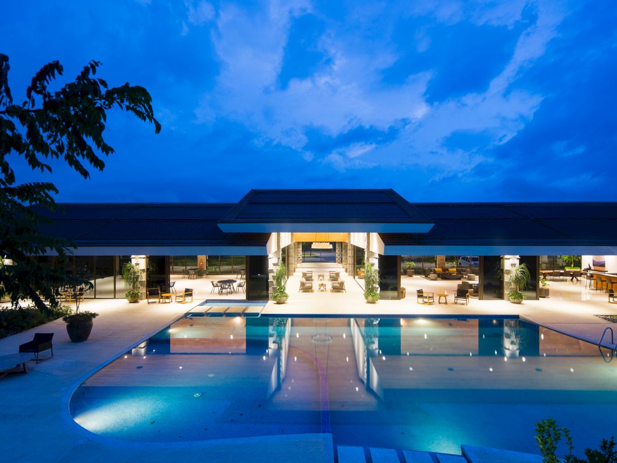 A luxurious building with large windows and a brightly lit interior overlooks a swimming pool under a twilight sky, surrounded by lounge chairs.