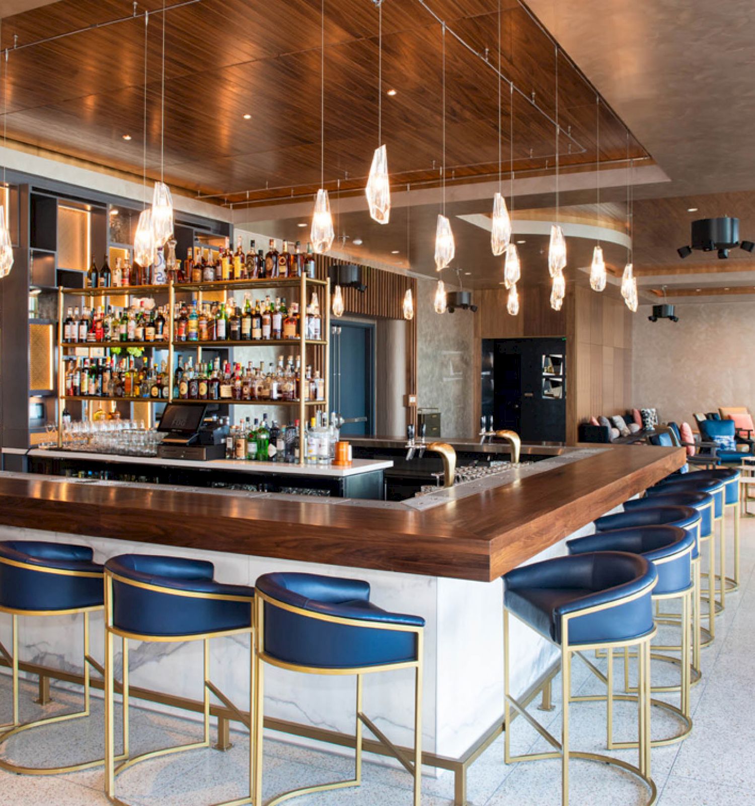 A modern bar area with a wooden countertop, blue and gold bar stools, hanging lights, and a well-stocked liquor shelf, alongside seating areas.
