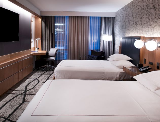 A modern hotel room with two neatly made beds, a wall-mounted TV, a work desk, and large windows with curtains, conveying a clean and stylish look.