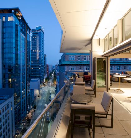 A modern cityscape at dusk featuring high-rise buildings and a well-lit balcony with tables and chairs, offering an urban view.