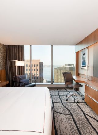 A modern hotel room with a large bed, desk, chair, flat-screen TV, patterned carpet, and window offering a city view with buildings and water.