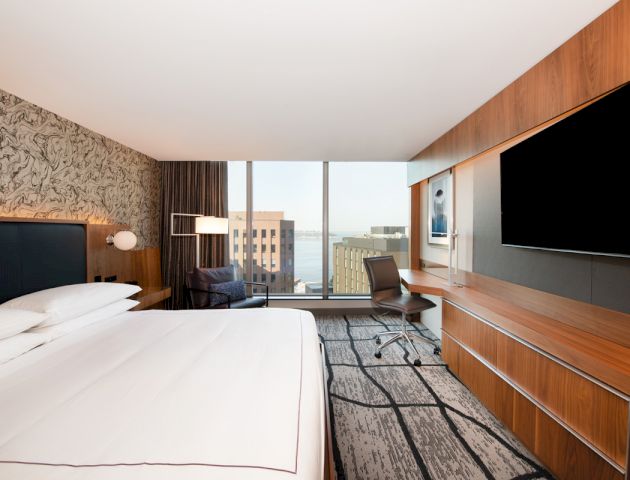 A modern hotel room with a large bed, desk, chair, flat-screen TV, patterned carpet, and window offering a city view with buildings and water.