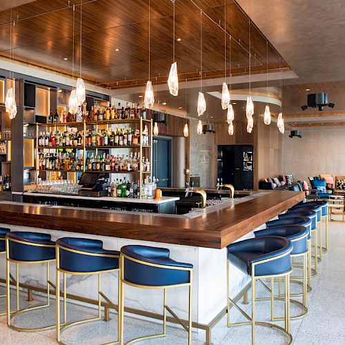 The image shows a modern bar with a wooden counter, bar stools with blue seats, a fully stocked bar, hanging lights, and a seating area in the background.