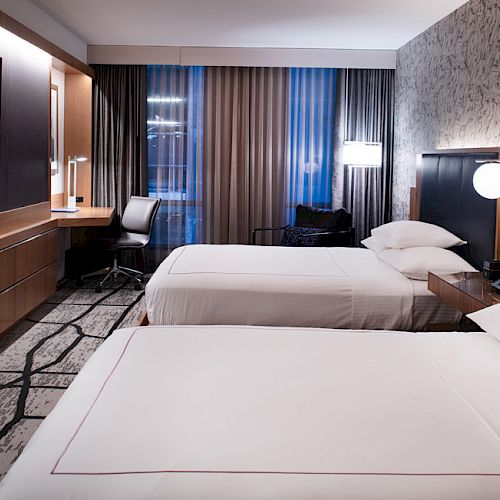 The image shows a modern hotel room with two neatly made beds, a desk with a chair, a mounted TV, and stylish decor with wall art and lighting fixtures.