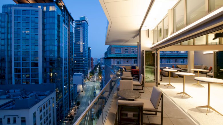 A modern cityscape is viewed from a stylishly furnished balcony, featuring high-rise buildings, outdoor seating, and ambient evening lighting.