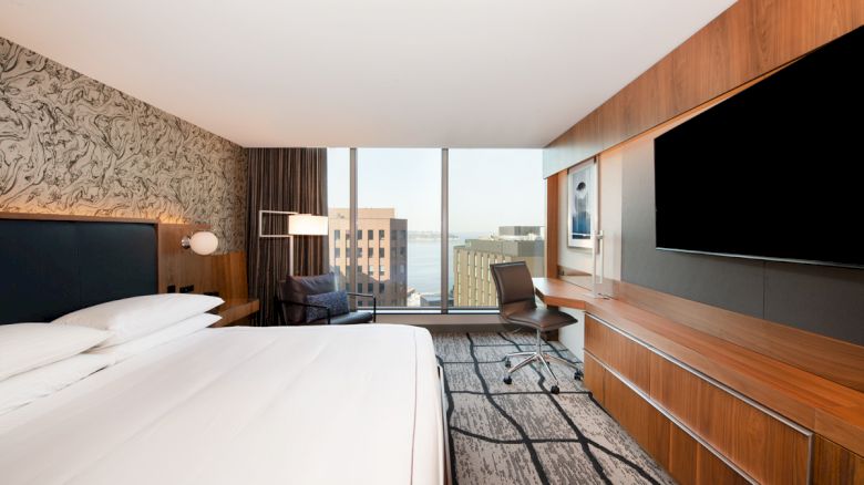 A modern hotel room with a large bed, desk, chair, TV, and a view of city buildings and water through floor-to-ceiling windows.