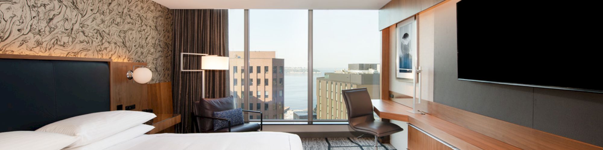 A modern hotel room with a large bed, wall-mounted TV, desk, chair, and a view of city buildings from a large window.